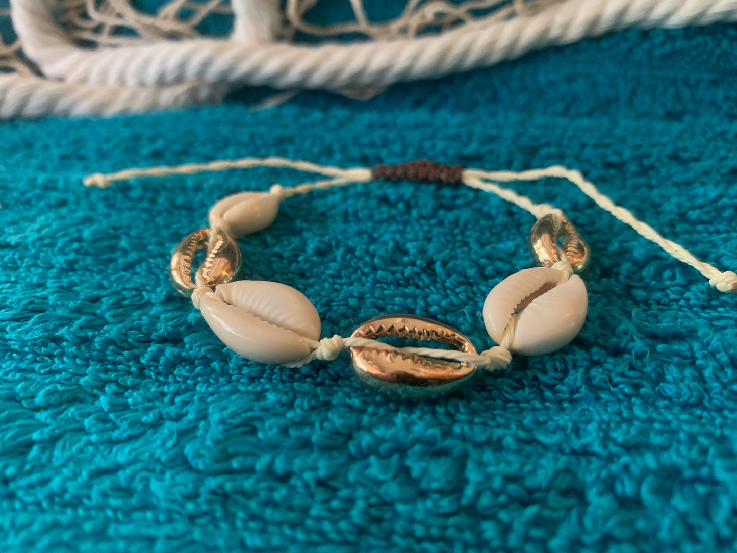 Pictured is a woven drawstring bracelet made with gold and regular cowrie shell beads. The bracelet is set on a towel-textured blue surface, with some netting and nautical rope blurred in the background