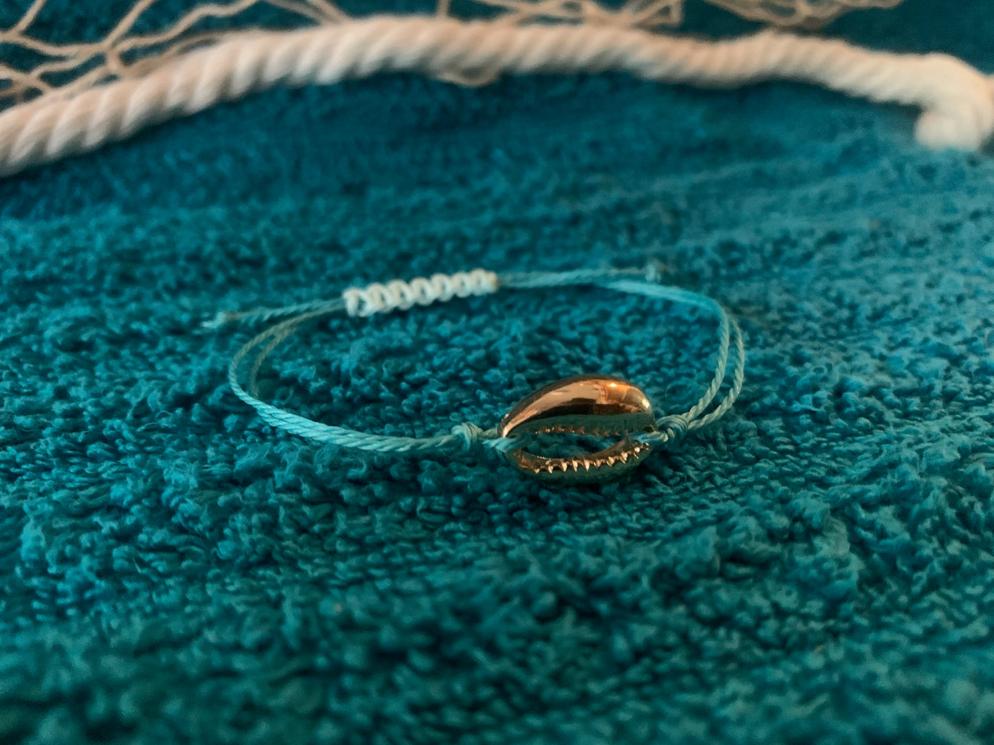 Bracelet with a gold cowrie shell charm on a textured teal towel-surface.