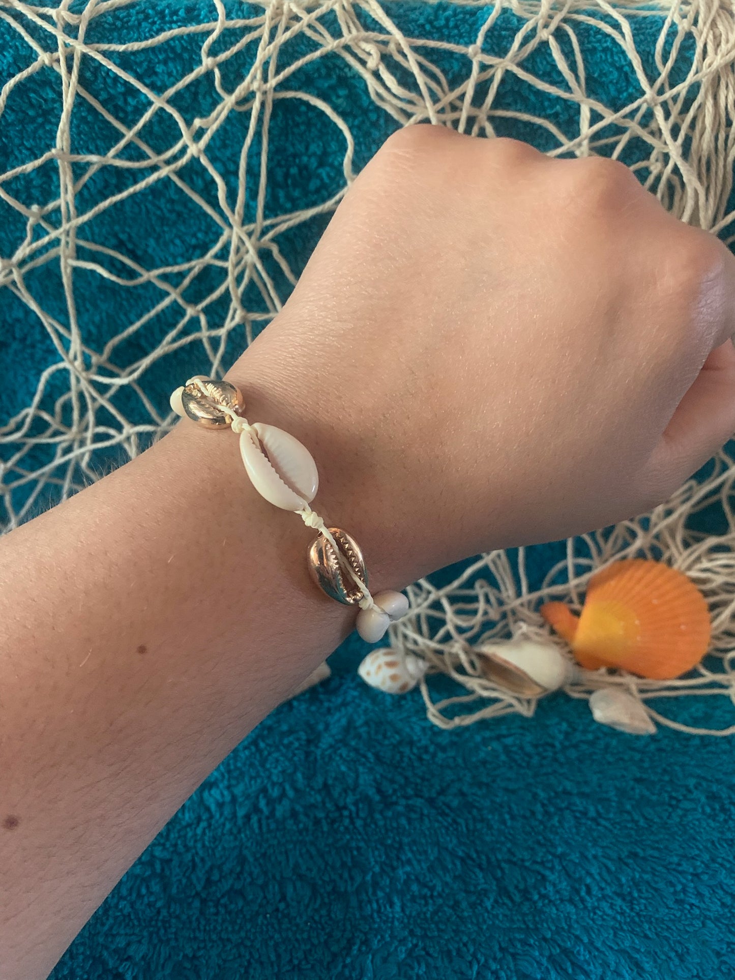 Pictured is a brown-skinned wrist wearing a woven drawstring bracelet made with gold and regular cowrie shell beads. The wrist and bracelet are held up in front of a towel-textured blue surface, with some netting, nautical rope, and seashells slightly blurred in the background.