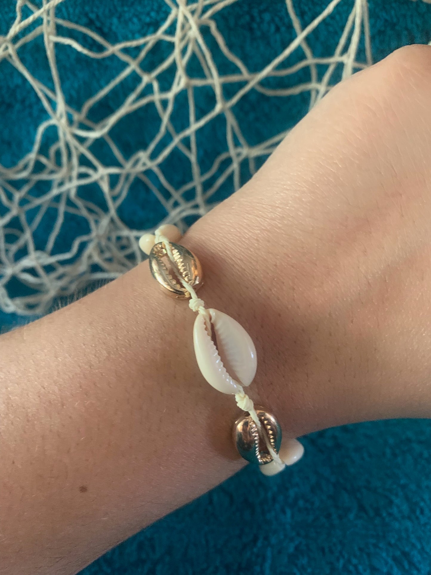 Pictured is a brown-skinned wrist wearing a woven drawstring bracelet made with gold and regular cowrie shell beads. The wrist and bracelet are held up in front of a towel-textured blue surface, with some netting and nautical rope blurred in the background