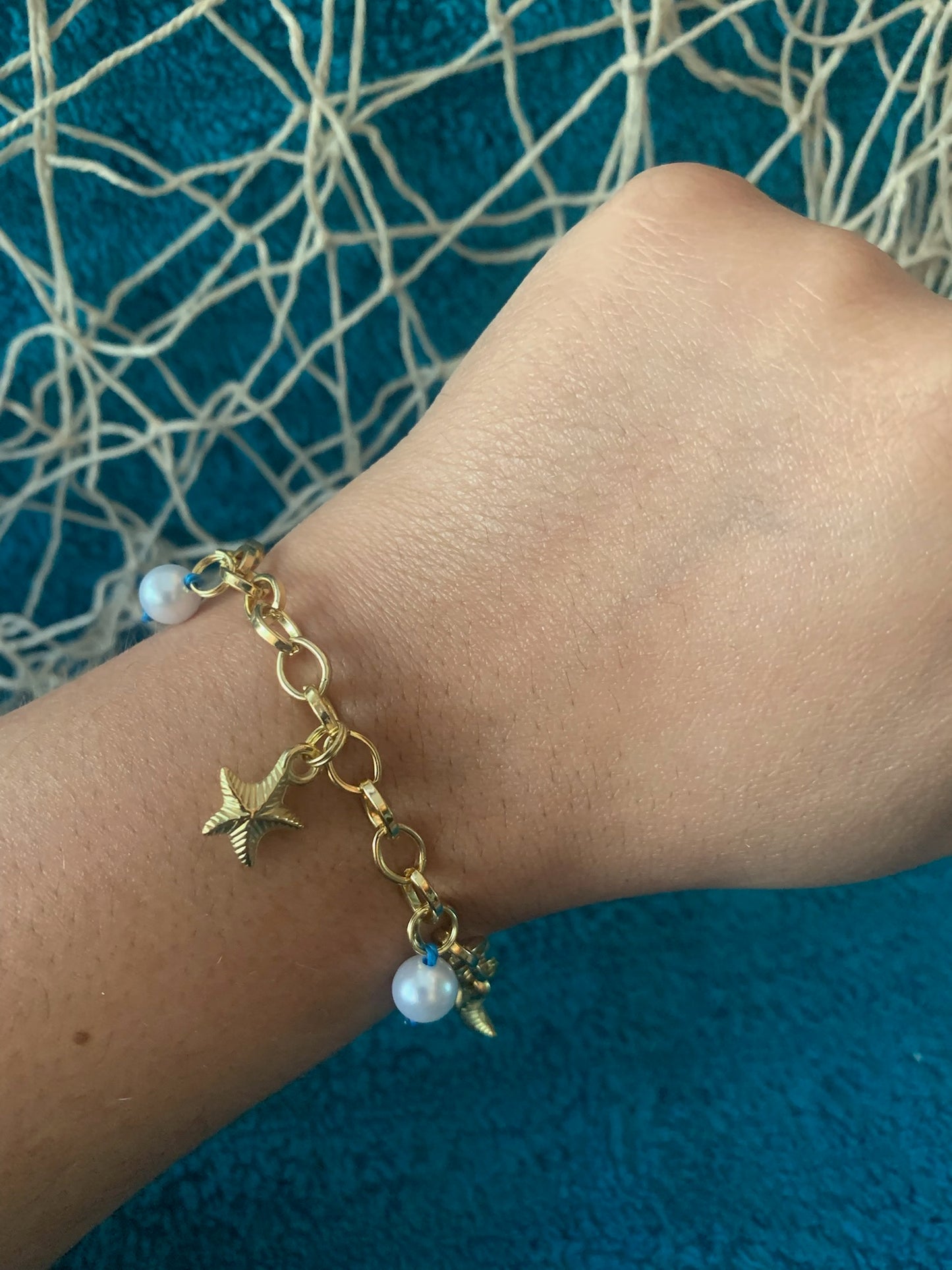 Gold bracelet with star and pearl charms on a wrist against a blue textured background