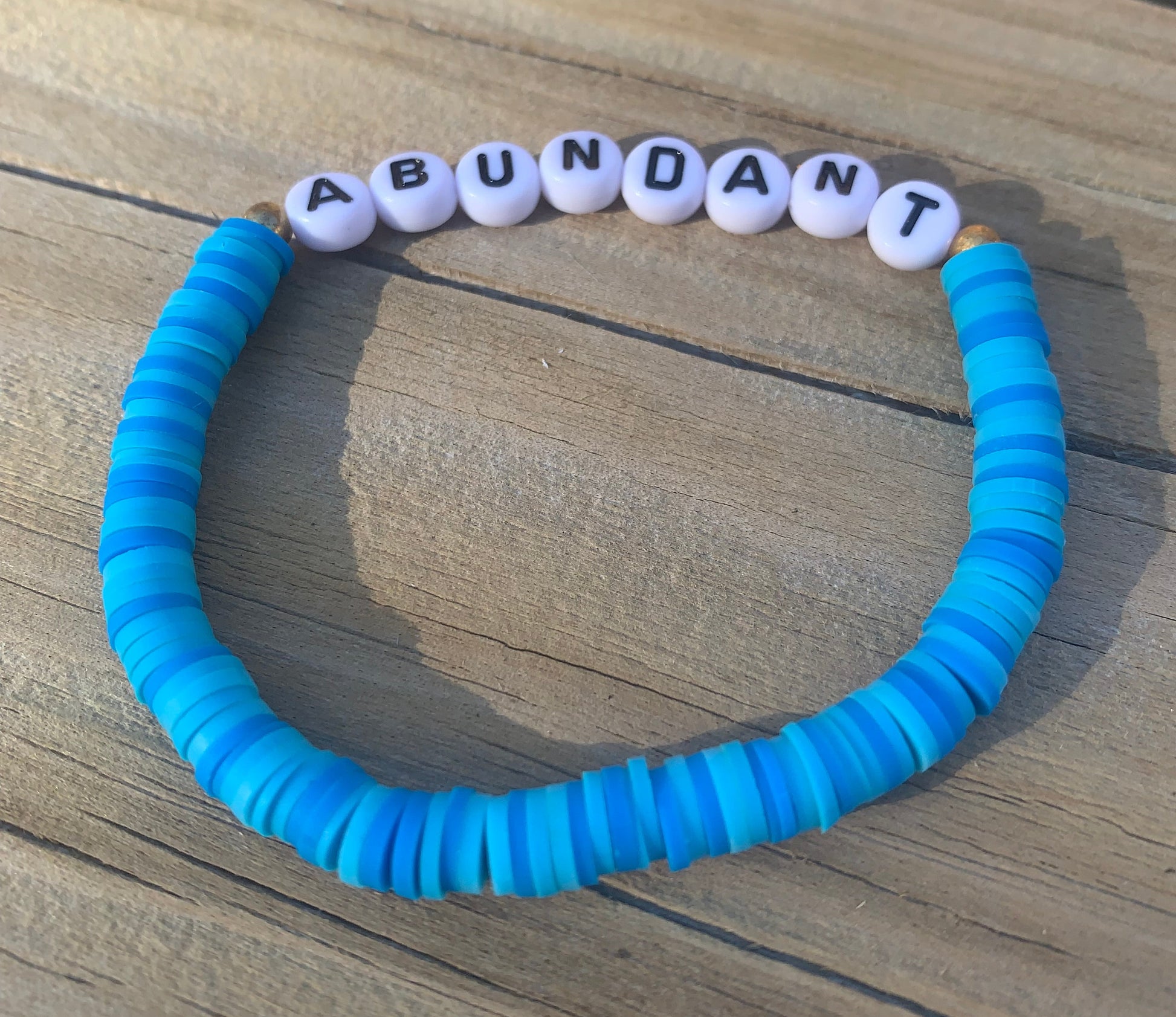 Blue textured bracelet with white beads spelling 'ABUNDANT' on a wooden surface