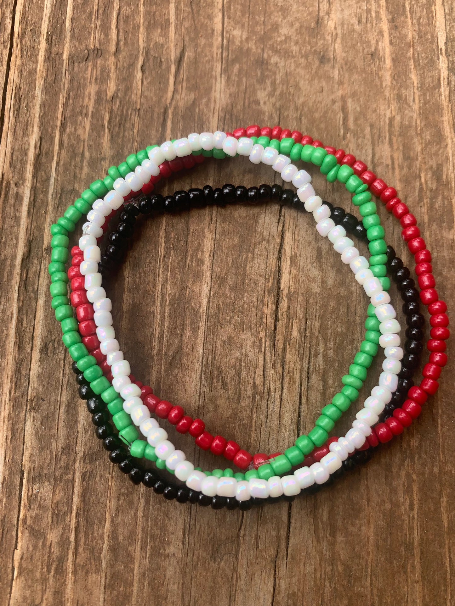 A set of four seed bead bracelets, one green, one red, one black, and one white, all stack atop each other in a bunch, on a distressed wood background.