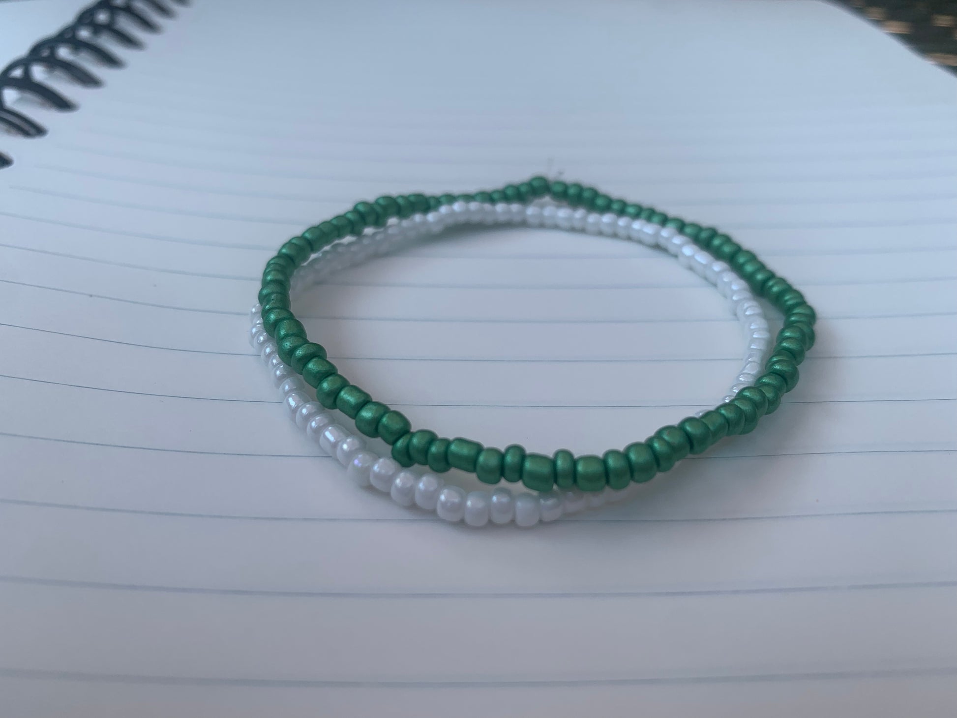 A Kelly-green seed bead bracelet is stacked atop a white seed bead bracelet, on a blue lined white paper notebook.