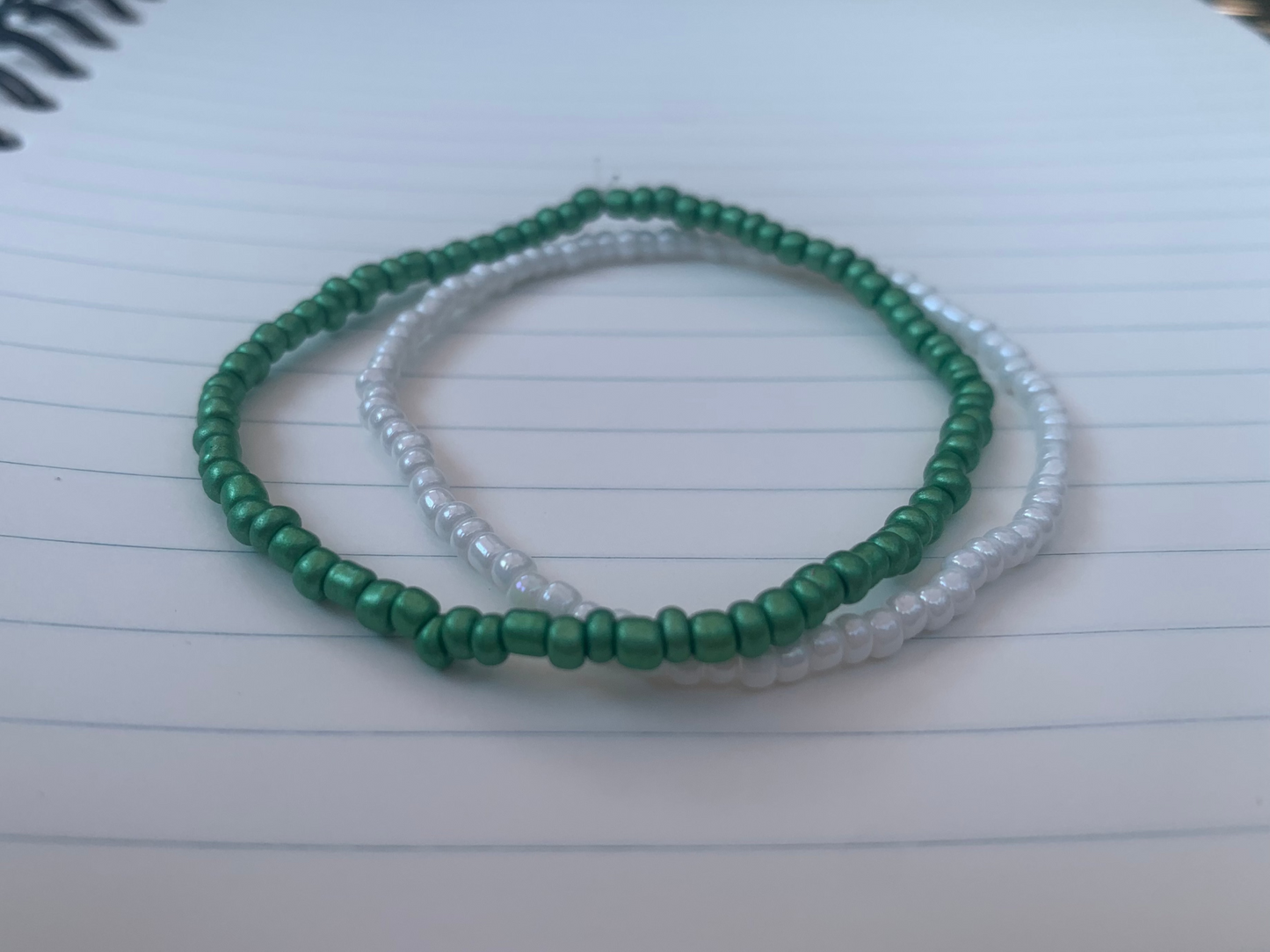 A Kelly-green seed bead bracelet is stacked atop a white seed bead bracelet, on a blue lined white paper notebook.