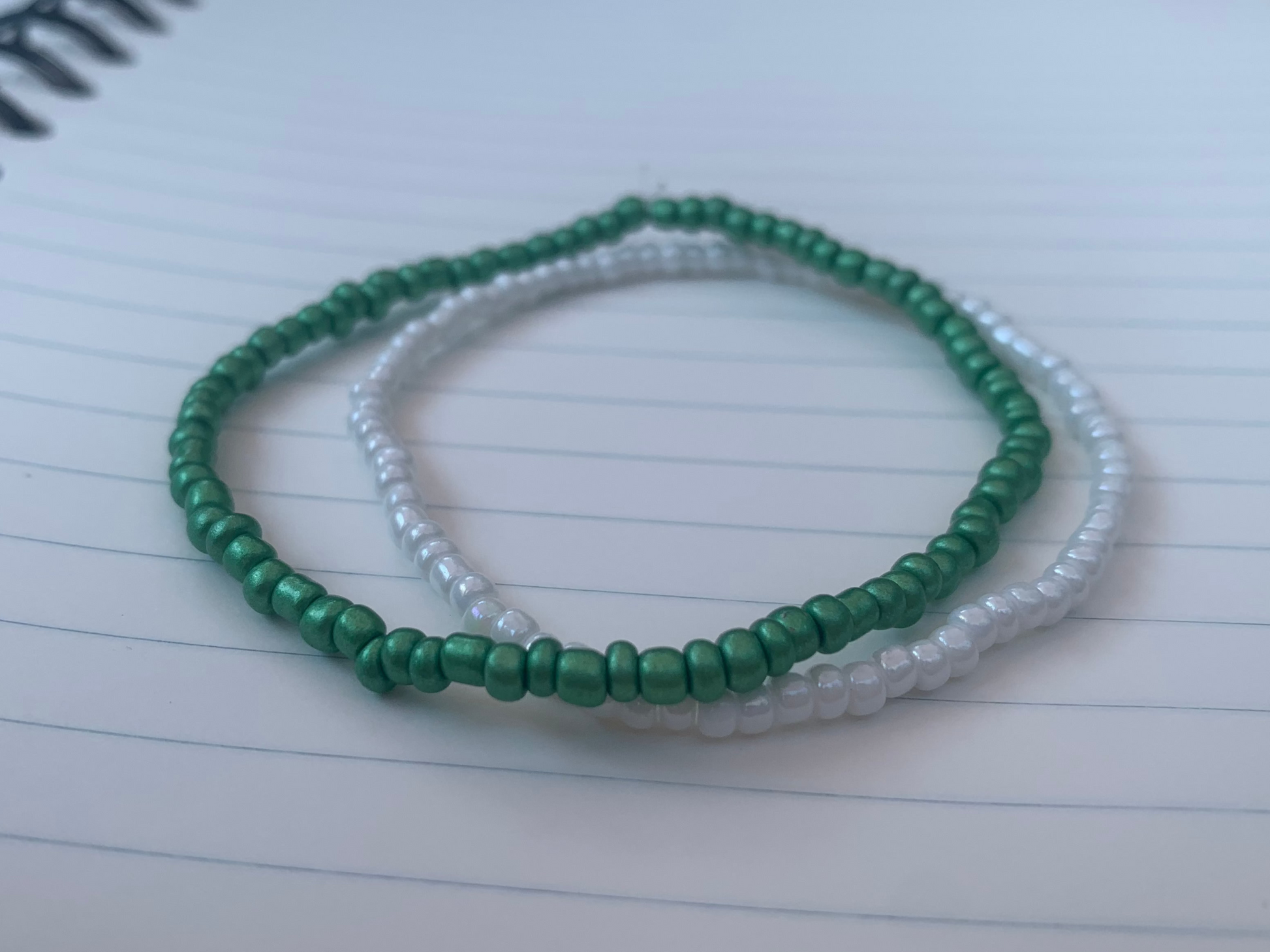 A Kelly-green seed bead bracelet is stacked atop a white seed bead bracelet, on a blue lined white paper notebook.