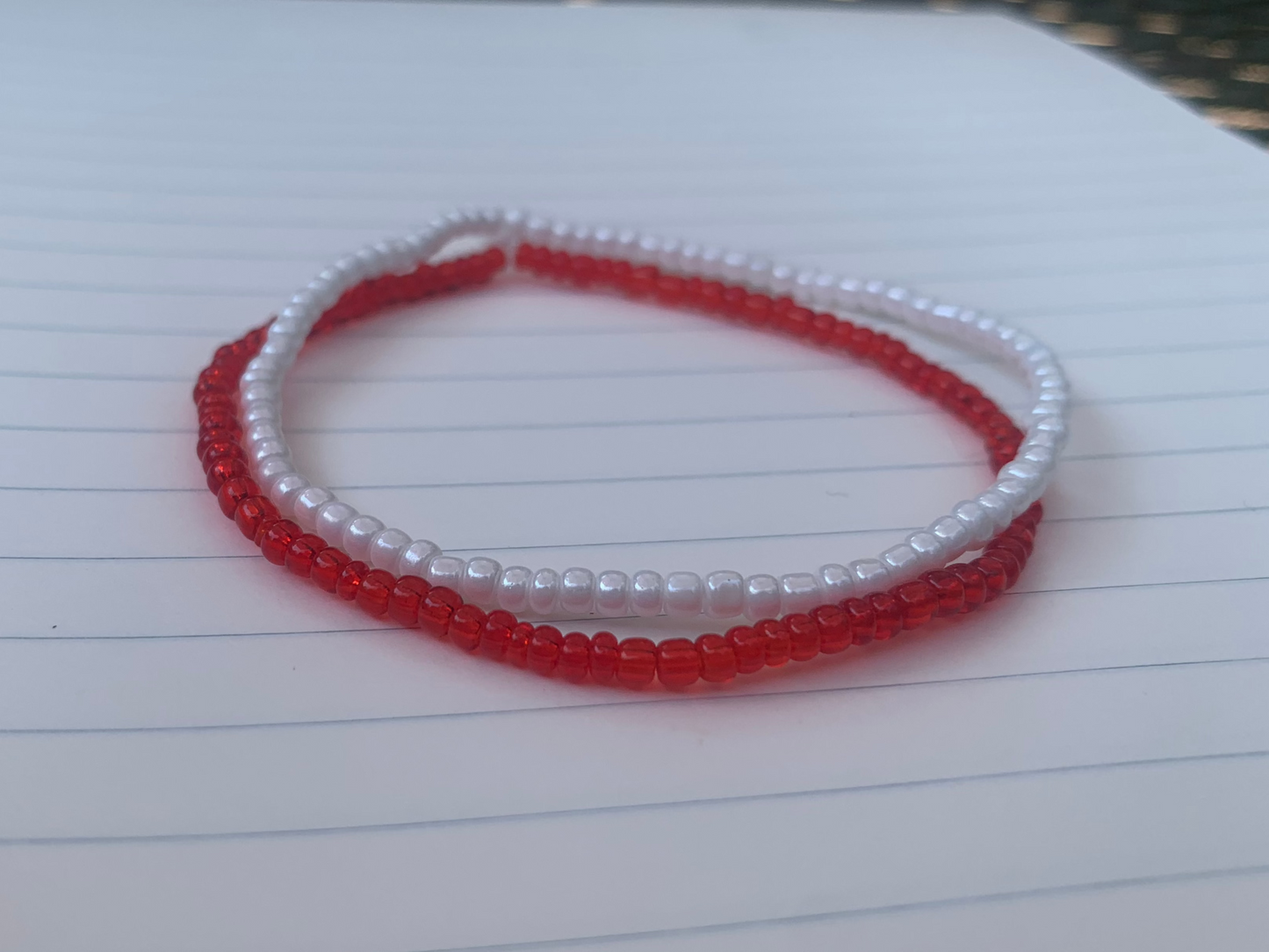 A white seed bead bracelet is stacked atop a translucent red seed bead bracelet, on a blue lined white paper notebook.