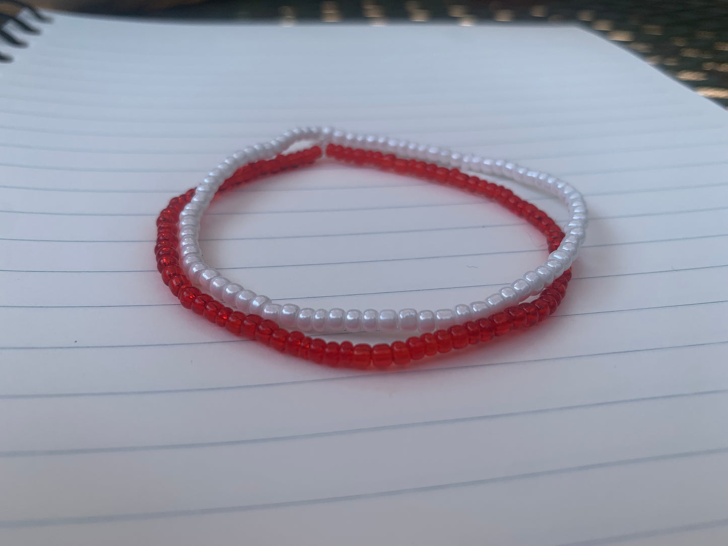 A white seed bead bracelet is stacked atop a translucent red seed bead bracelet, on a blue lined white paper notebook.