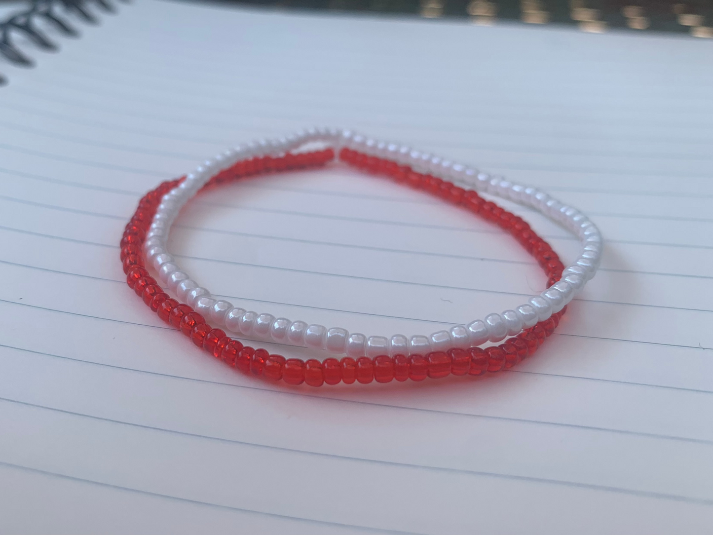 A white seed bead bracelet is stacked atop a translucent red seed bead bracelet, on a blue lined white paper notebook.