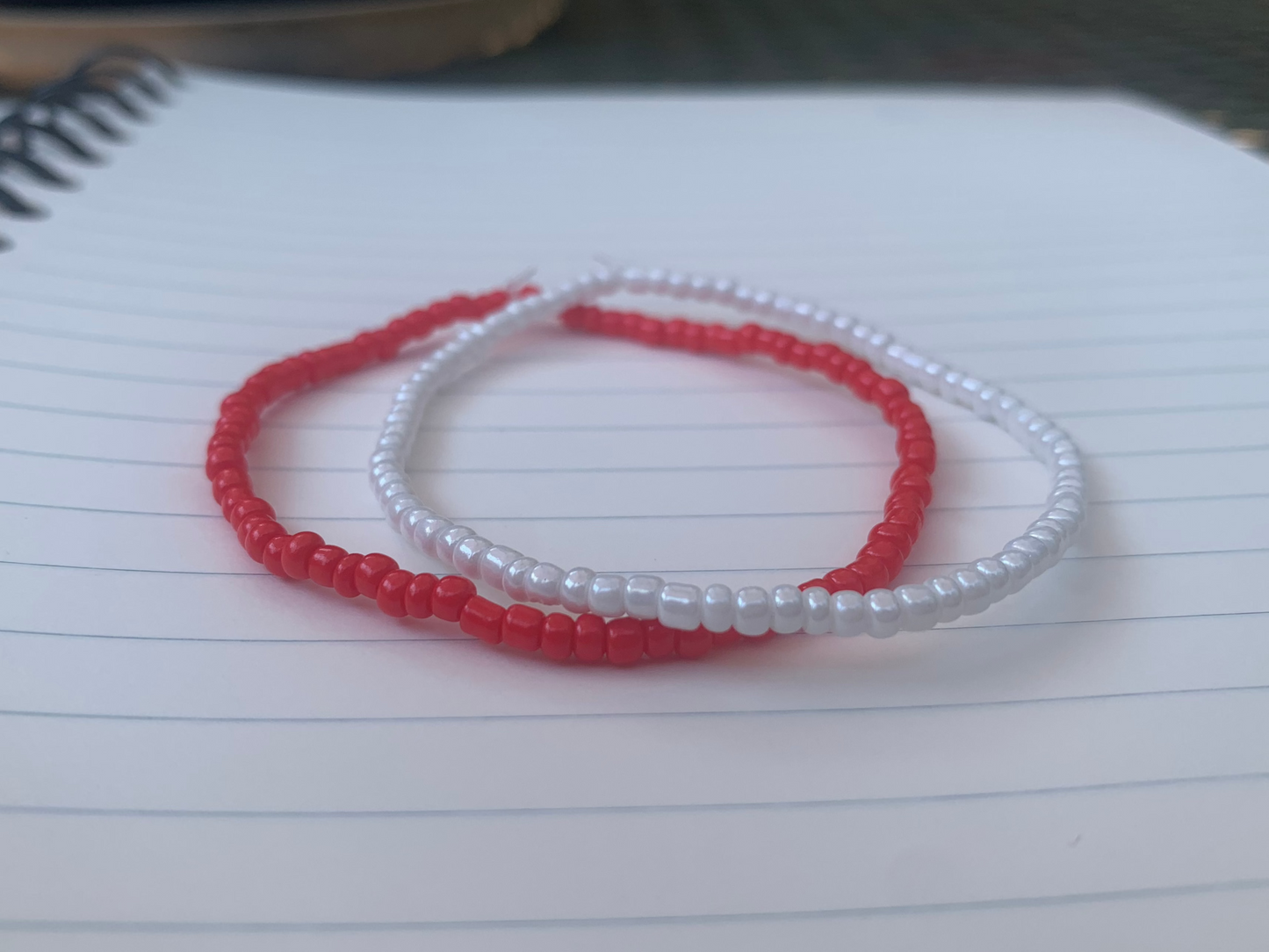  A vermillion red seed bead bracelet is stacked atop a white seed bead bracelet, on a blue lined white paper notebook.