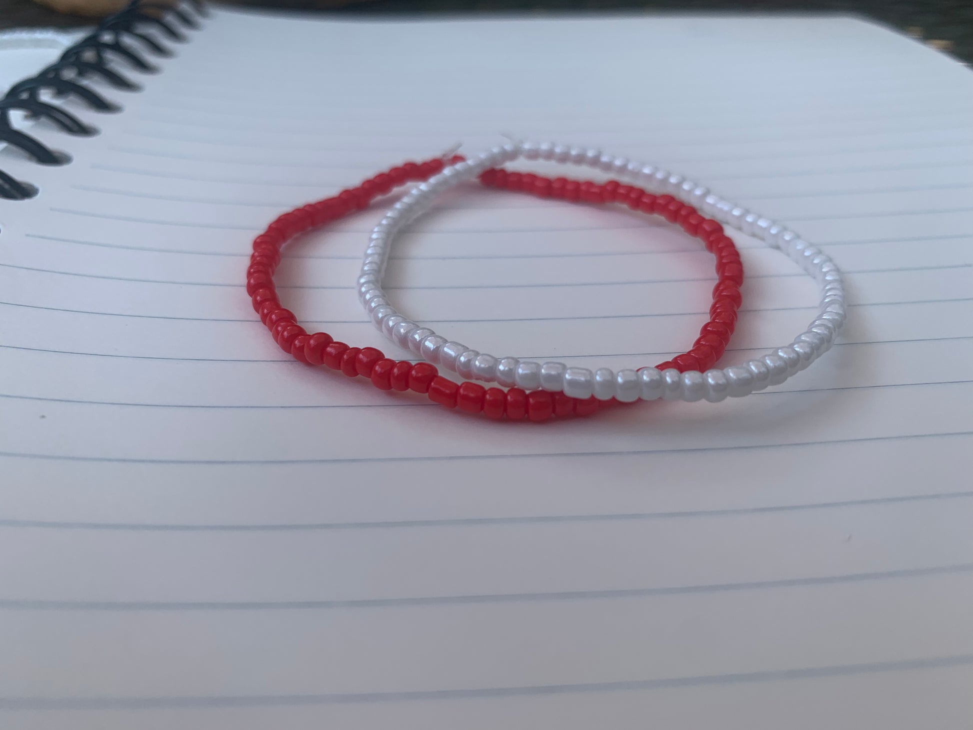  A vermillion red seed bead bracelet is stacked atop a white seed bead bracelet, on a blue lined white paper notebook.