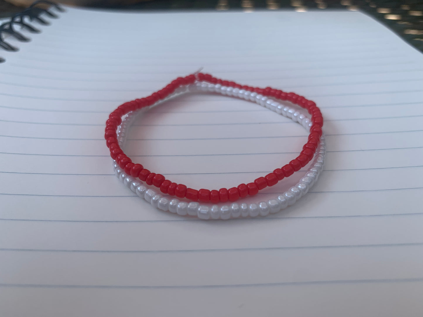  A vermillion red seed bead bracelet is stacked atop a white seed bead bracelet, on a blue lined white paper notebook.