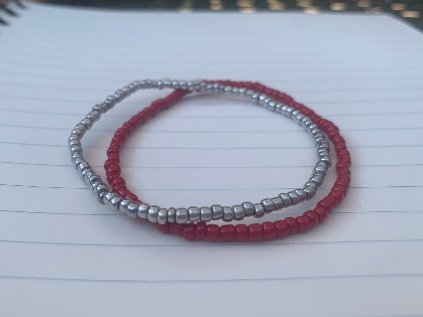 A silvery-grey seed bead bracelet is stacked atop a dark red seed bead bracelet, on a blue lined white paper notebook.