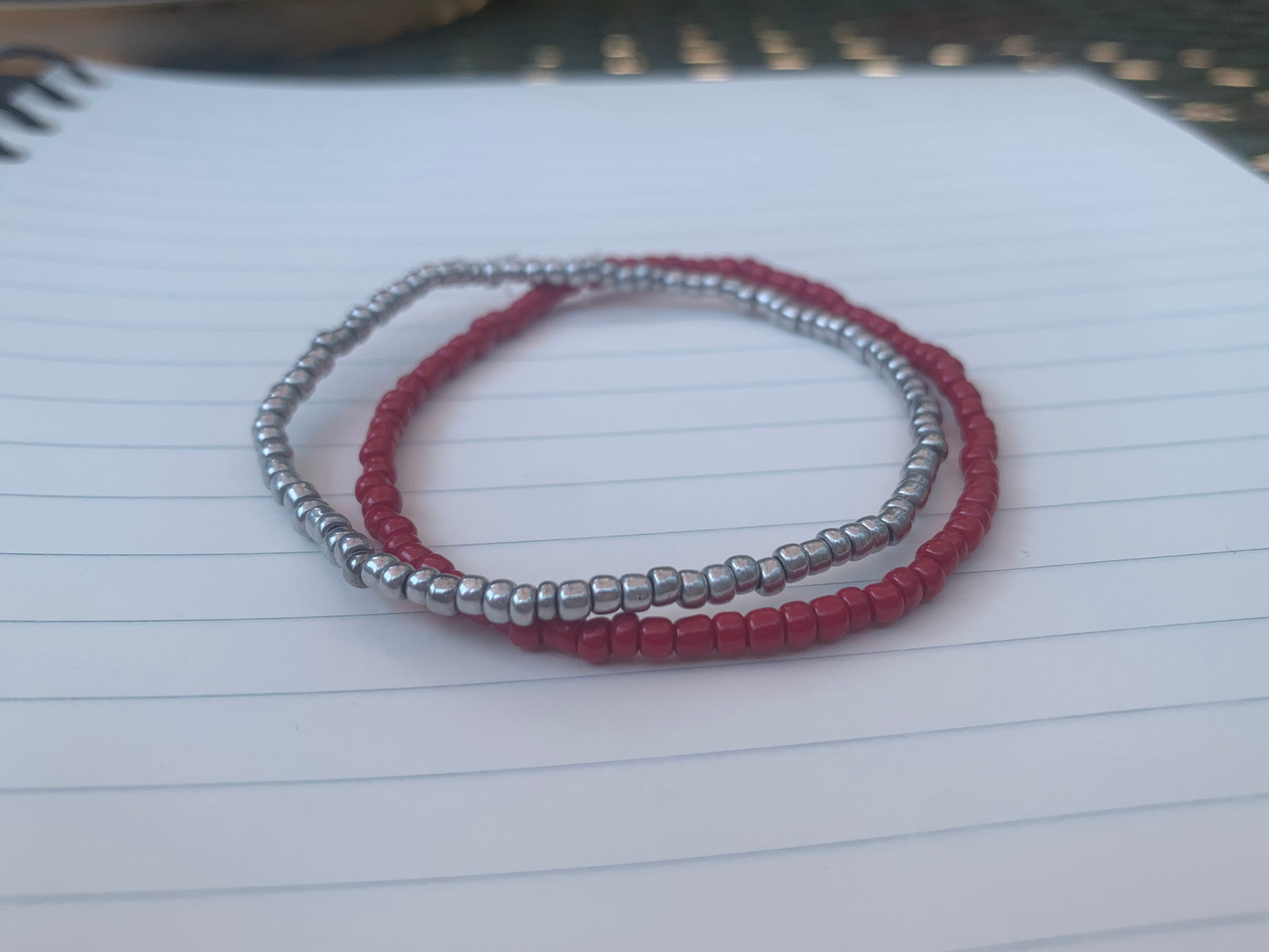 A silvery-grey seed bead bracelet is stacked atop a dark red seed bead bracelet, on a blue lined white paper notebook.