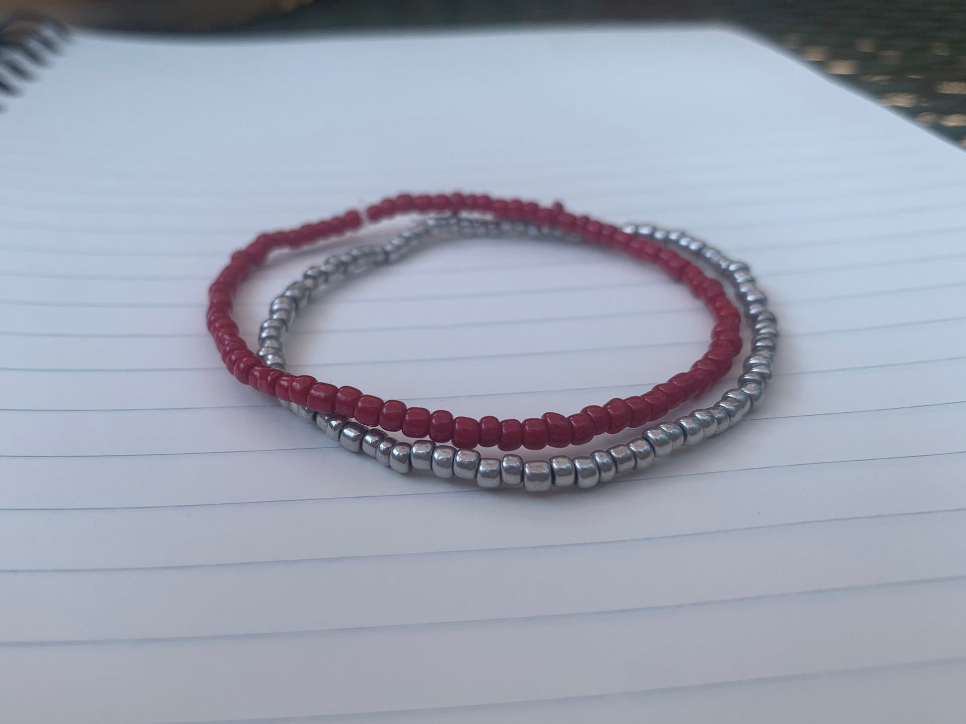 A dark red seed bead bracelet is stacked atop a silvery-grey seed bead bracelet, on a blue lined white paper notebook.