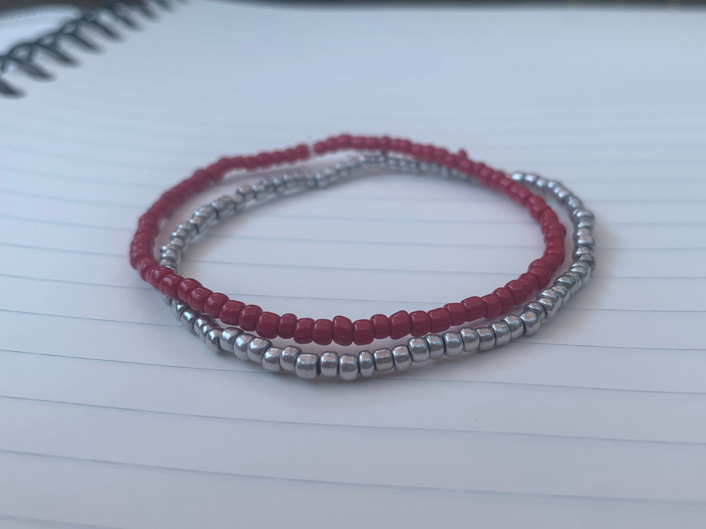 A dark red seed bead bracelet is stacked atop a silvery-grey seed bead bracelet, on a blue lined white paper notebook.