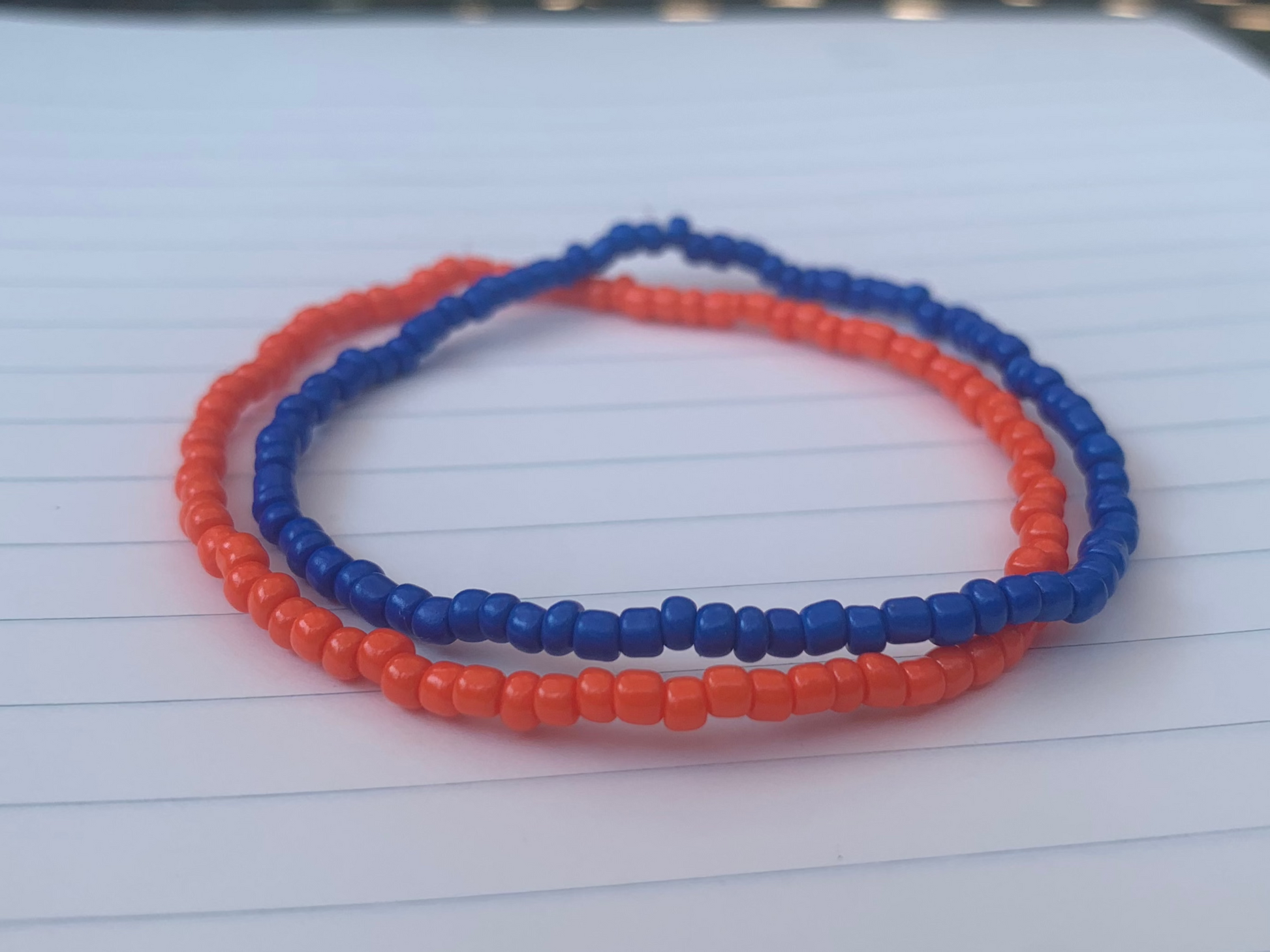 A dark blue seed bead bracelet is stacked atop an orange seed bead bracelet, on a blue lined white paper notebook. 