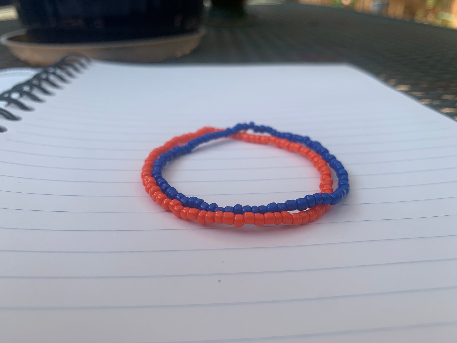 A dark blue seed bead bracelet is stacked atop an orange seed bead bracelet, on a blue lined white paper notebook. 