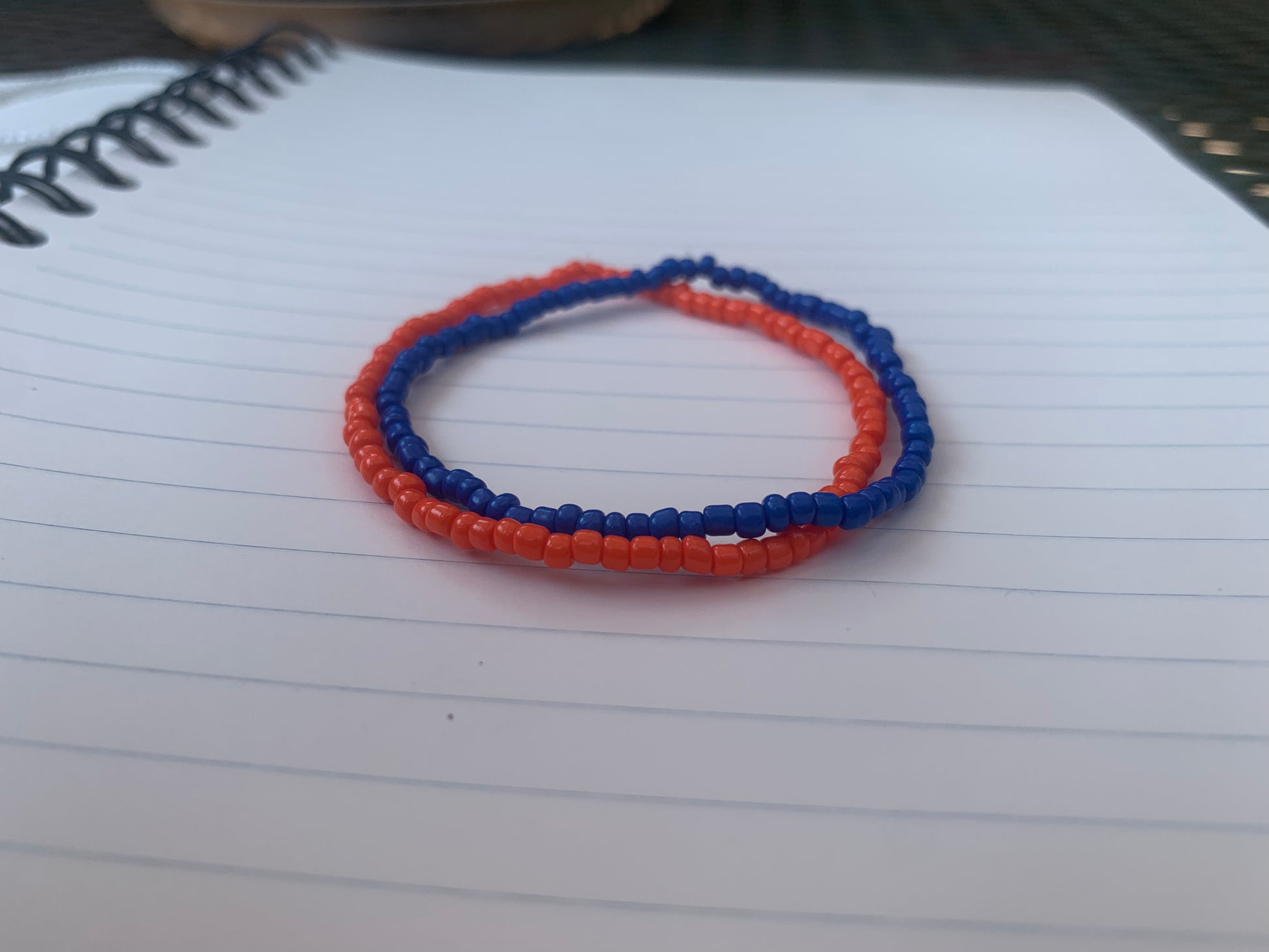 A dark blue seed bead bracelet is stacked atop an orange seed bead bracelet, on a blue lined white paper notebook. 