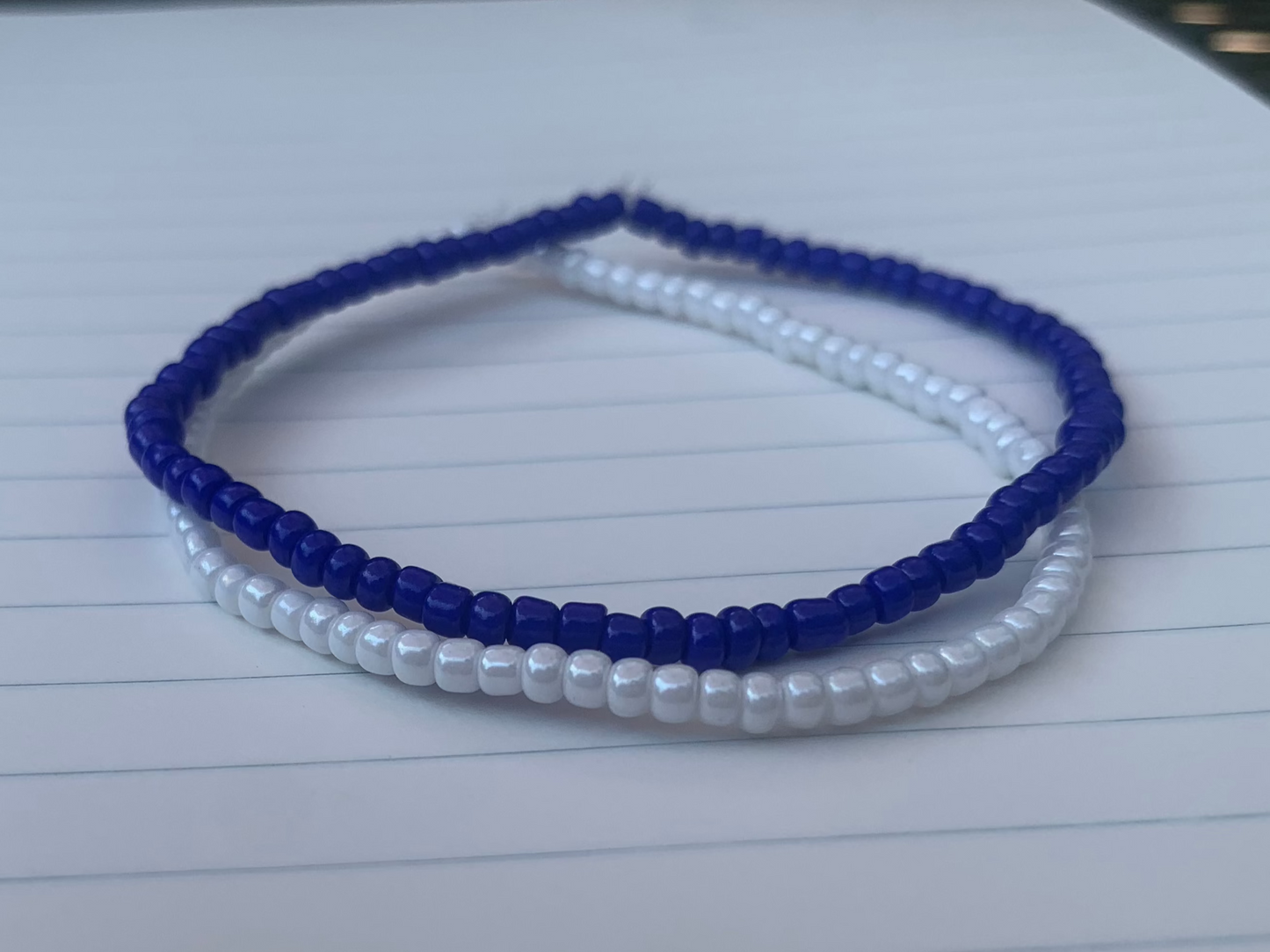 A dark blue seed bead bracelet is stacked atop a white seed bead bracelet, on blue lined white notebook paper.