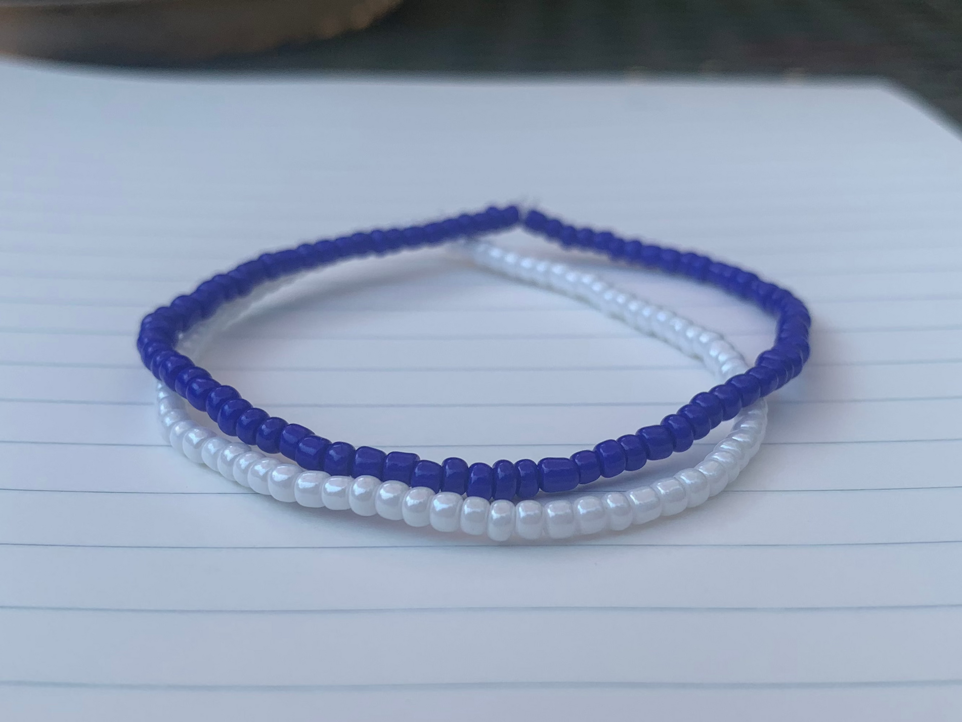 A dark blue seed bead bracelet is stacked atop a white seed bead bracelet, on blue lined white notebook paper.