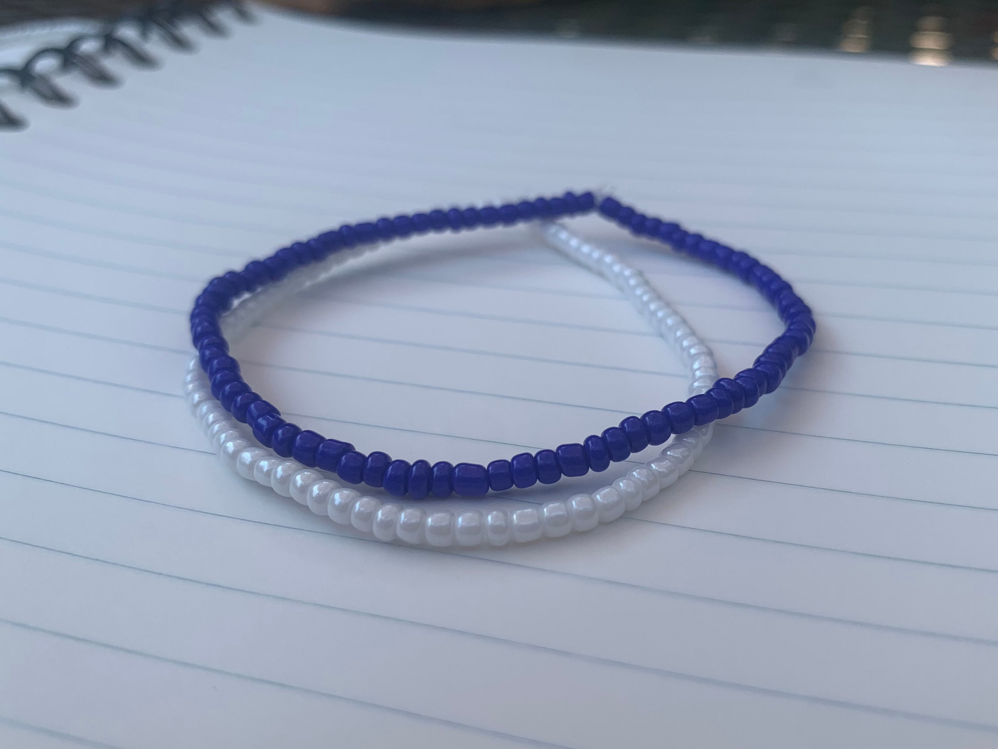 A dark blue seed bead bracelet is stacked atop a white seed bead bracelet, on blue lined white notebook paper.