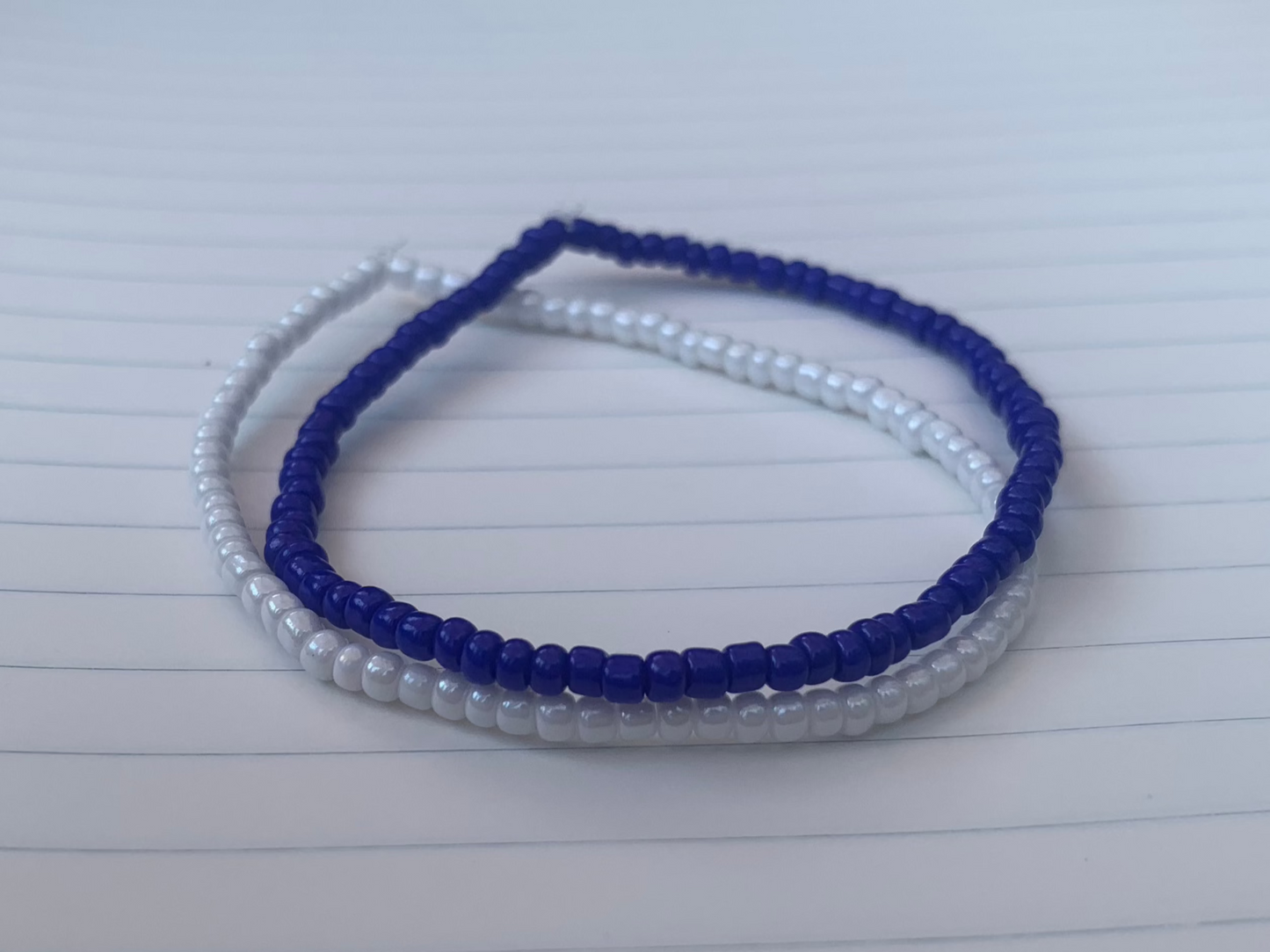 A dark blue seed bead bracelet is stacked atop a white seed bead bracelet, on blue lined white notebook paper.