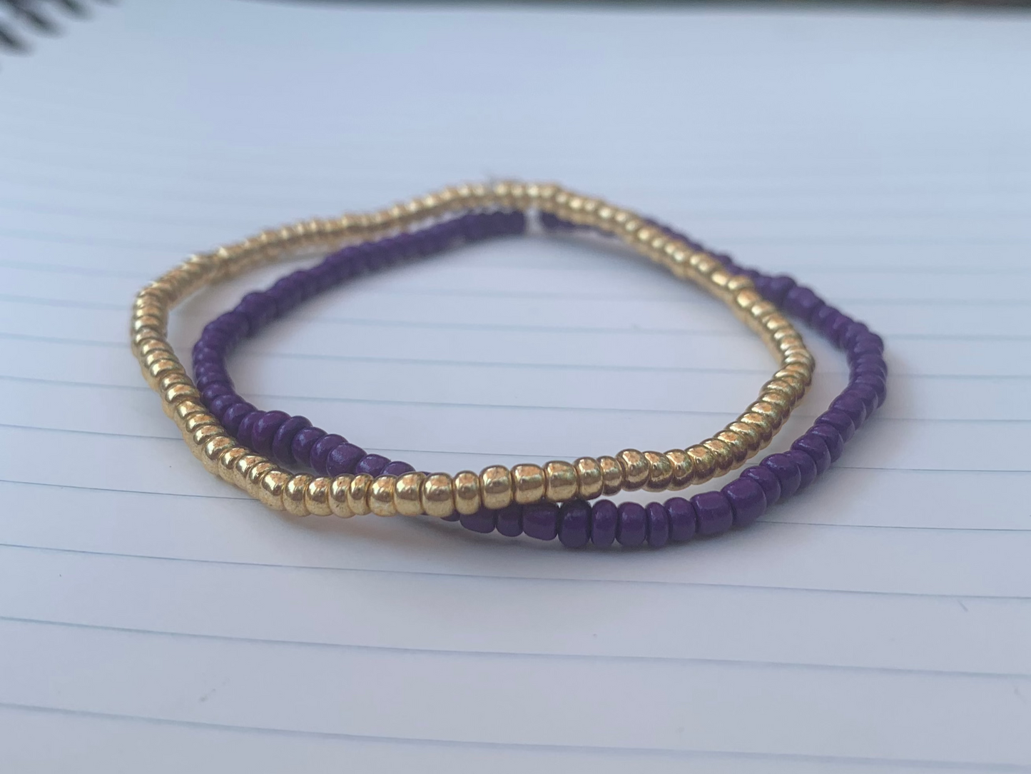 A gold seed bead bracelet is stacked atop a dark purple seed bead bracelet, on a blue lined white paper notebook.