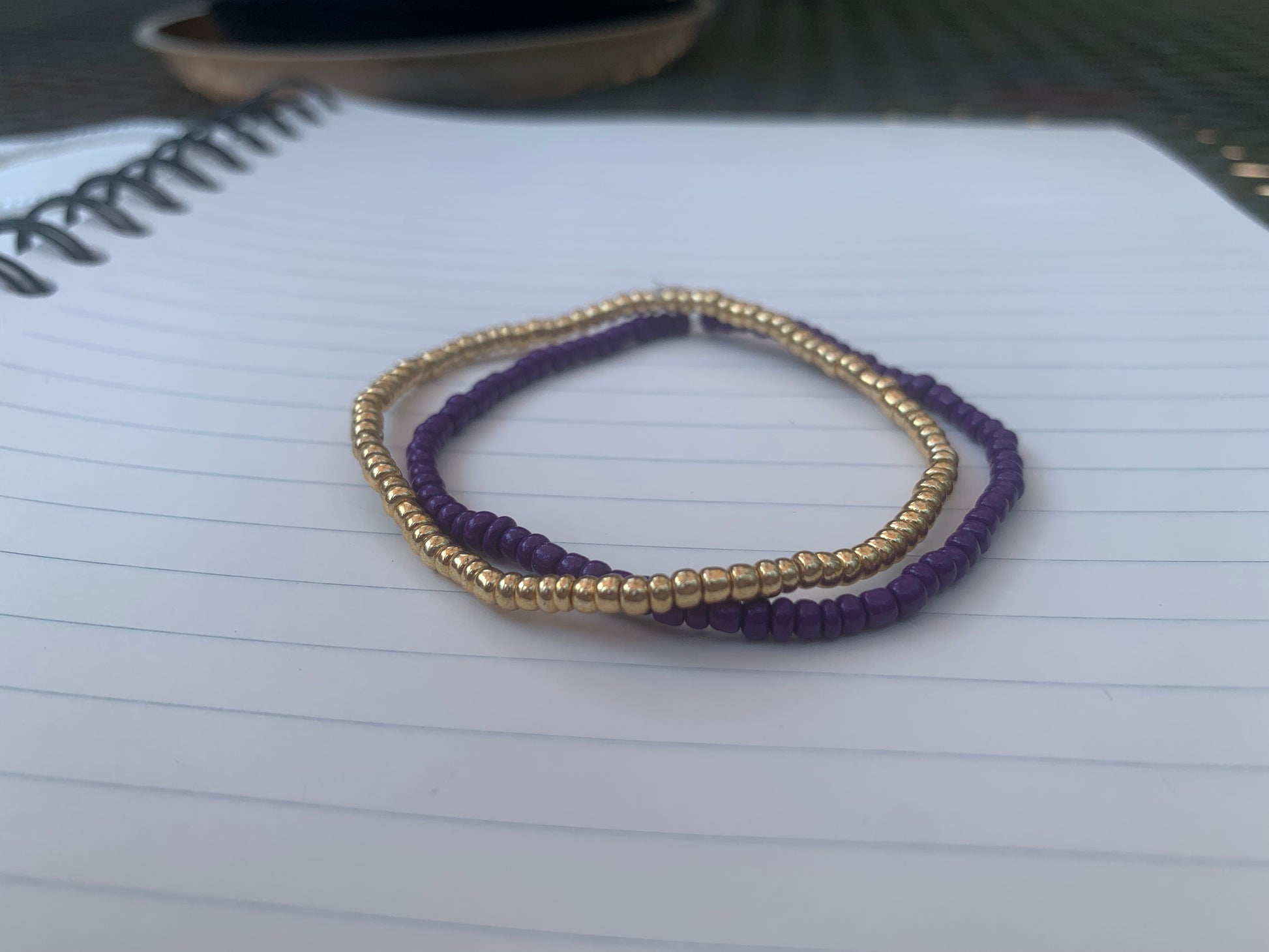 A gold seed bead bracelet is stacked atop a dark purple seed bead bracelet, on a blue lined white paper notebook.