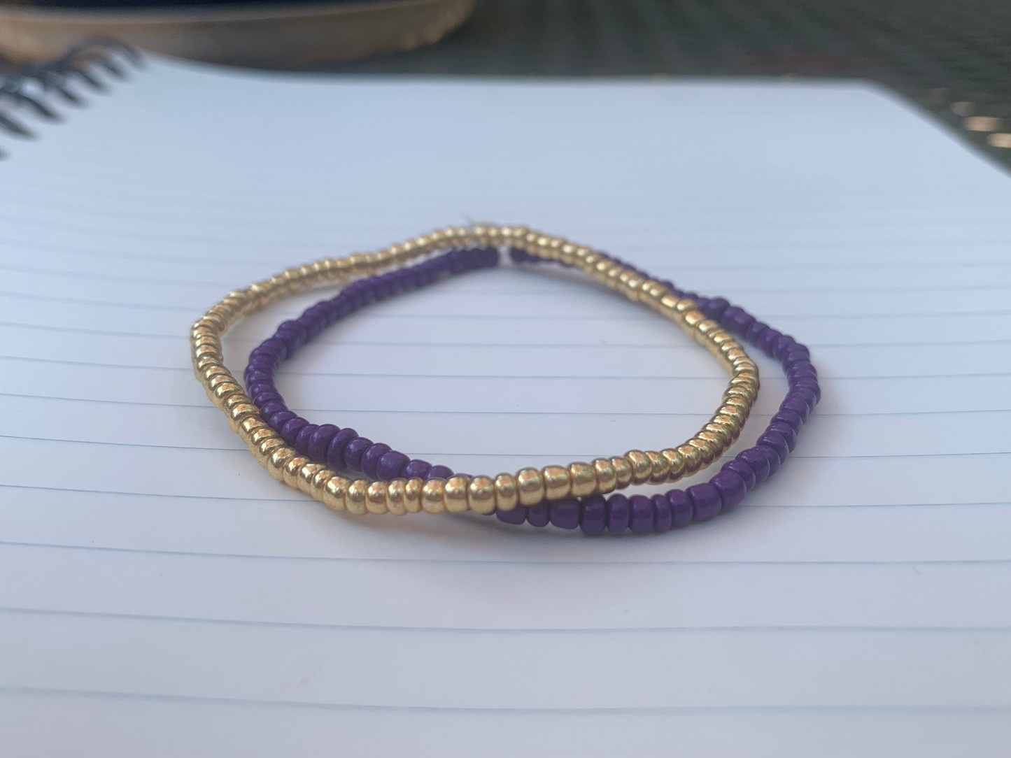 A gold seed bead bracelet is stacked atop a dark purple seed bead bracelet, on a blue lined white paper notebook.