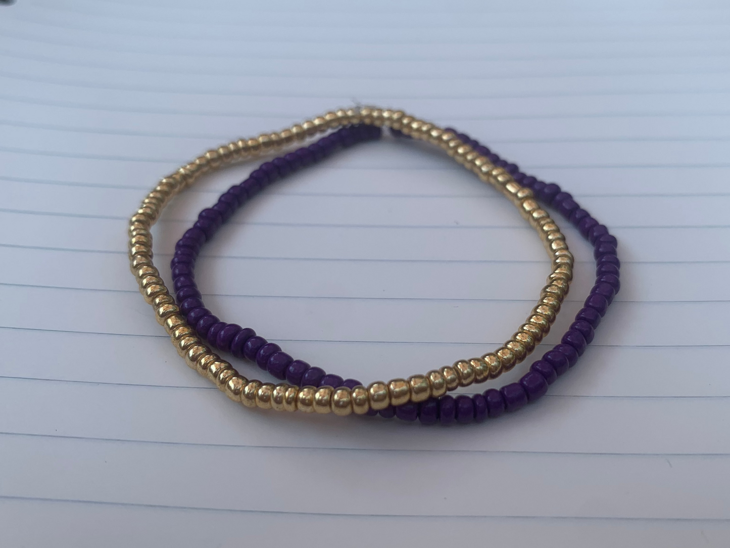 A gold seed bead bracelet is stacked atop a dark purple seed bead bracelet, on a blue lined white paper notebook.