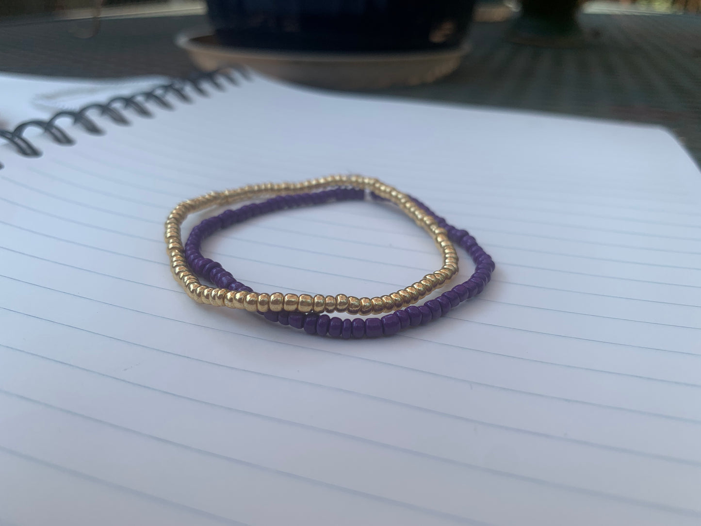 A gold seed bead bracelet is stacked atop a dark purple seed bead bracelet, on a blue lined white paper notebook.