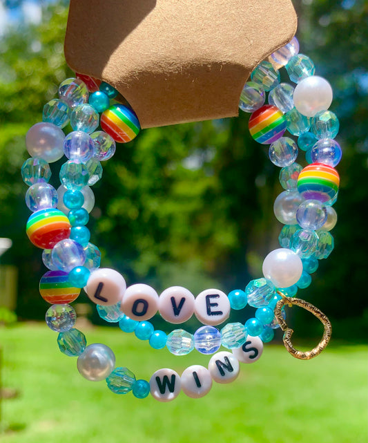 Beaded bracelet with blue, pearl, and rainbow striped beads and a gold heart charm on a natural background. Black and white letter beads spell out "LOVE WINS"
