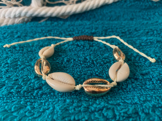 Pictured is a woven drawstring bracelet made with gold and regular cowrie shell beads. The bracelet is set on a towel-textured blue surface, with some netting and nautical rope blurred in the background