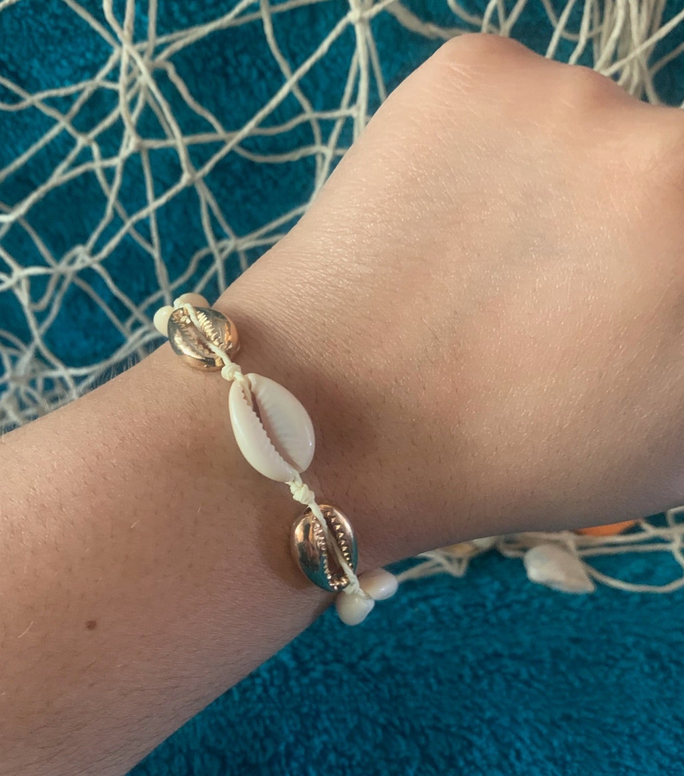 Pictured is a brown-skinned wrist wearing a woven drawstring bracelet made with gold and regular cowrie shell beads. The wrist and bracelet are held up in front of a towel-textured blue surface, with some netting and nautical rope blurred in the background.