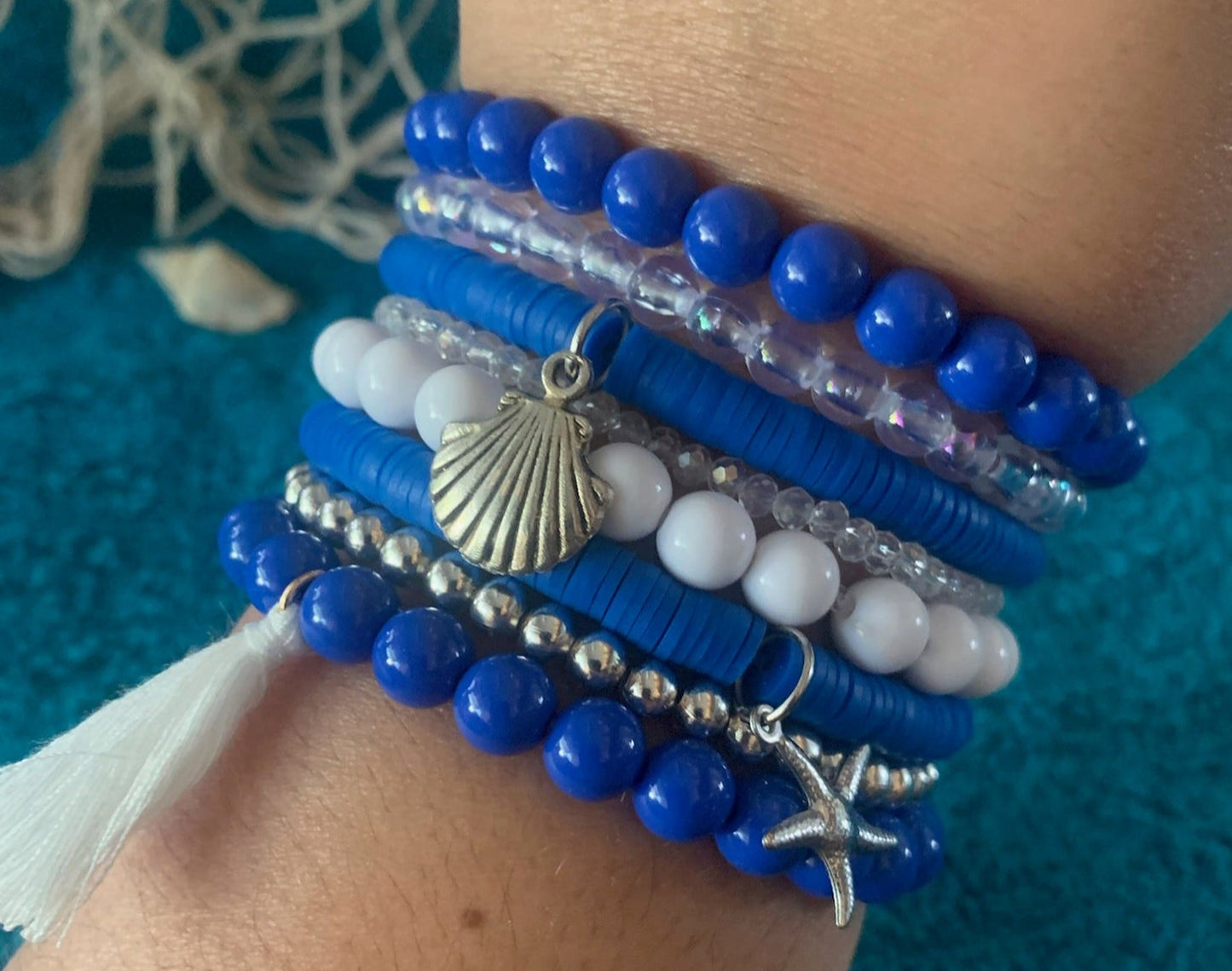 Close-up of a wrist wearing multiple blue and white beaded bracelets with charms on a textured blue background.