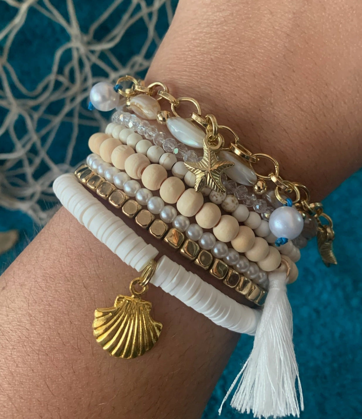Close-up of a wrist wearing multiple beaded bracelets with shell and tassel charms against a textured blue background.
